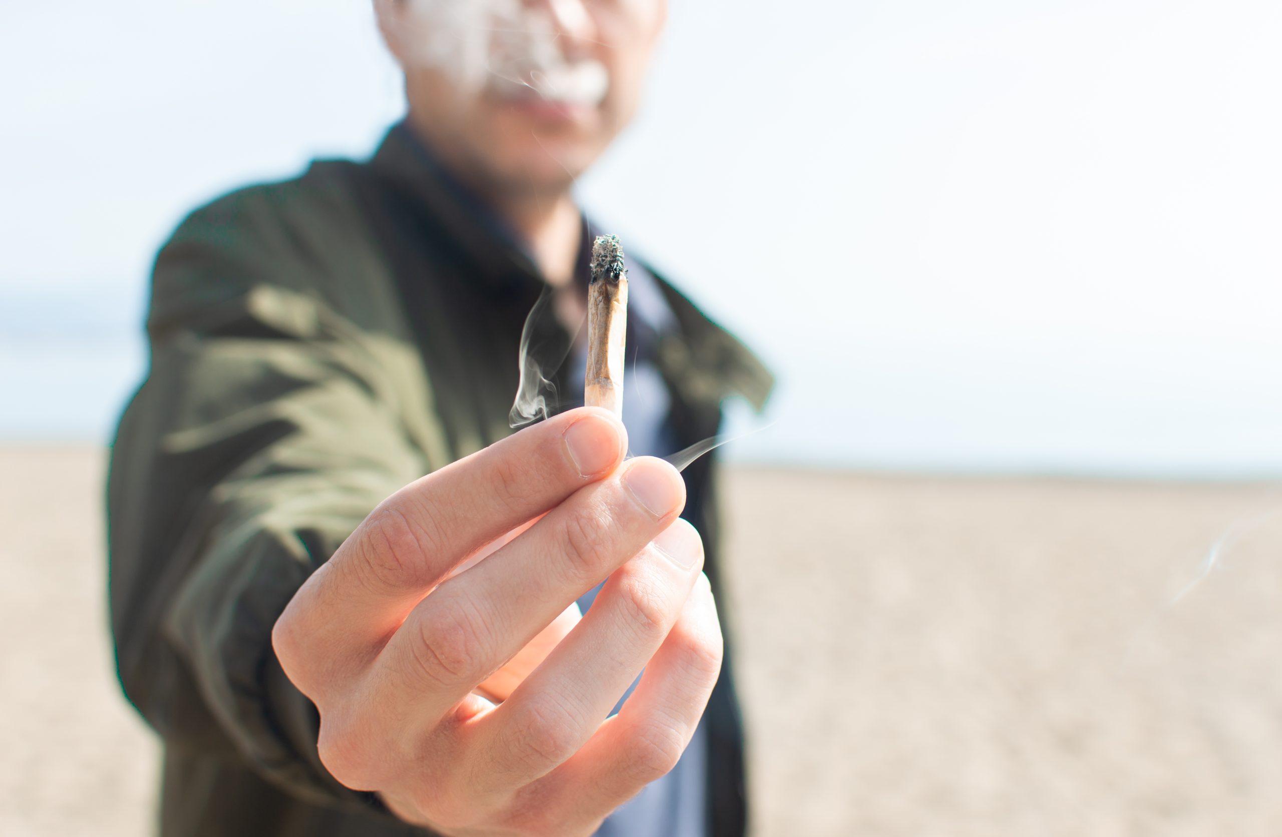 Young man holding a lit marijuana joint while smoking on the beach.